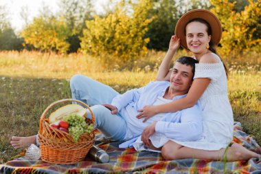 Çimenli bir alanda, ağaçlı bir battaniyenin üzerinde oturan bir çift. Güneşli bir öğleden sonra meyve dolu bir sepetin yanında gülümser ve dinlenirler..