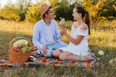 Erkek ve kadın tarlada battaniyede oturur, sepet ve çiçeklerin meyvelerini paylaşır, doğada güneşli bir günde birlikte güler ve gülerler..
