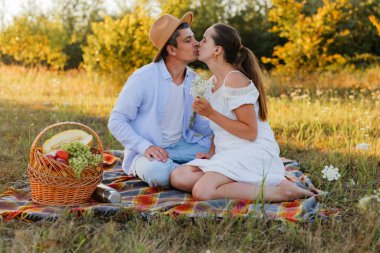 Bir tarlada battaniyeye oturup öpüşen iki insan. Meyve ve çiçeklerle dolu bir piknik sepetleri var, dışarıda sıcak bir günün tadını çıkarıyorlar..