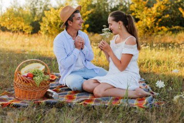 Genç bir çift, bir tarlada bir battaniyeye oturup gülüşmeler ve kahkahalar atıyor. Ellerinde meyve ve çiçeklerle dolu bir piknik sepeti var..