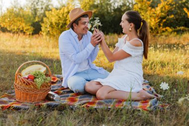 İki insan bir tarlada battaniyeye oturup birbirlerine gülümsüyor. Güneşli bir günde ellerinde çiçekler ve meyve dolu bir piknik sepeti olur..