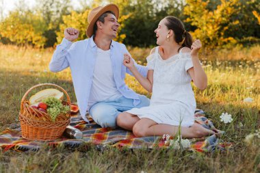 İki insan bir tarlada battaniyeye oturmuş, birlikte gülüyor ve gülümsüyor. Orada bir sepet dolusu meyveler vardır. Gün ışığı sahnede parlar.