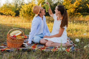İki kişi çimenli bir tarlada bir battaniyeye oturmuş, gülümsüyor ve üzümleri paylaşıyor. Etrafı meyveyle dolu bir piknik sepetiyle çevrili..