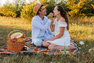 Bir erkek, güneşli bir tarlada piknik yaparken bir kadını bir parça meyveyle besler. Bir sepet yiyecekle battaniyenin üzerinde otururken gülümser ve gülerler..