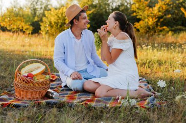 Bir çift tarlada bir battaniyeye oturup meyve paylaşıyor. Doğayla ve sıcak güneş ışığıyla çevriliyken gülümserler ve birbirlerine girerler..