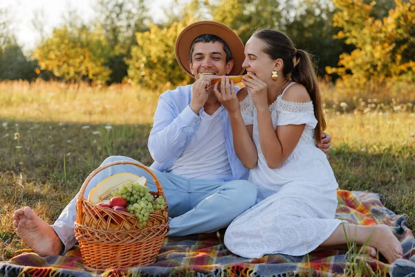 İki kişi çimenli bir alanda battaniyeye oturup sepetten meyve yiyor. Güneşli bir günde bir anı paylaşırken gülümserler..