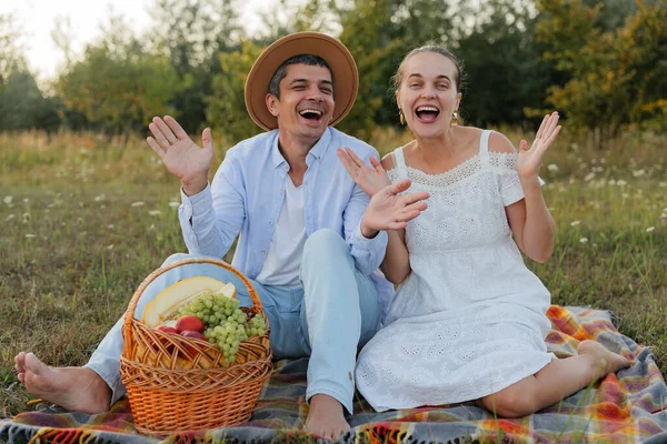 İki kişi çimenli bir tarlada bir battaniyeye oturup bir sepet meyveyle gülümseyerek ellerini sallıyor. Güneş arka planda batıyor..
