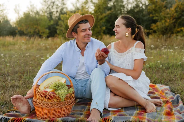 Bir çift sıcak akşam ışığında bir tarlada battaniyeye oturup sepetteki meyveleri paylaşır ve birbirlerine gülümserler..
