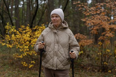 Bir kadın elinde bastonla ormanda duruyor. Örtülü bir ceket ve şapka giyiyor. Ağaçlar sonbahar mevsimini gösteren sarı yapraklar sergiliyor..