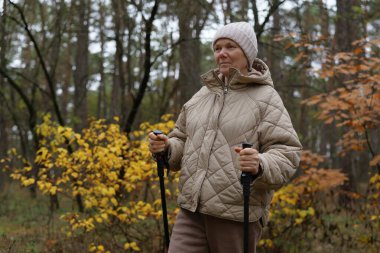 Bir kadın elinde bastonla ormanda duruyor. Örtülü bir ceket ve şapka giyiyor. # Sonbahar yaprakları sararmış # # Etrafını sarmış yürümekten zevk alırken #.