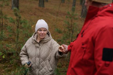 Bej ceketli bir kadın, ağaçlarla çevrili bir şekilde ormanda dikilirken kırmızı ceketli birinin elini tutmak için uzanır..
