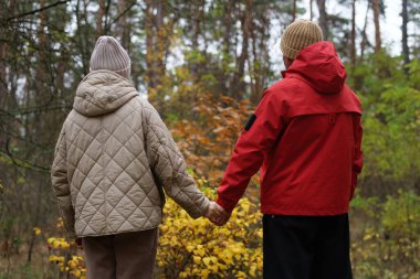 İki insan ormanda birlikte yürürken el ele tutuşuyorlar. Ağaçların sonbahar yaprakları var. Hava serin ve atmosfer rahat..