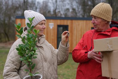 Bir insan bir tencerede küçük bir ağaç tutarken diğeri konuşur ve bir kutu tutar. Bulutlu bir havada, ahşap bir binanın yakınındaki bir bahçede duruyorlar..