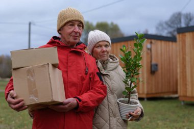 Yaşlı bir çift küçük ahşap evlerin önünde duruyor. Kadın saksıda bitki taşırken adam kutuyu tutuyor. Gökyüzü bulutlu ve gri.