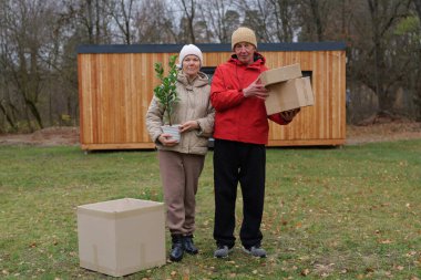 Arkalarında ahşap bir yapı olan bir parkta çimlerin üzerinde iki kişi duruyor. Biri saksıyı tutuyor, diğeri de iki kutu tutuyor. Sonbahar geldi..