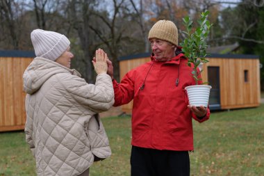 Biri küçük bir saksı ağacı tutarken iki yaşlı birey etkileşime girer. Dışarıda duruyorlar, ahşap bahçe yapılarıyla çevrili, serin bir günde..