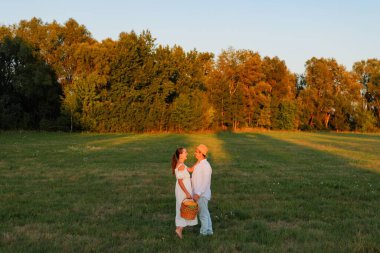 İki kişi el ele tutuşurken bir tarlada duruyor. Ağaçların yakınındalar ve bir sepetleri var. Güneş batıyor, sıcak bir atmosfer yaratıyor..