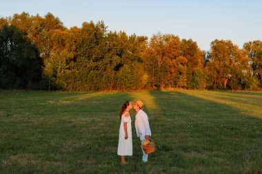 Gün batımında iki kişi çim tarlasında yüz yüze durur. Birbirlerine doğal bir şekilde gülümserken meyve dolu bir sepet tutarlar..