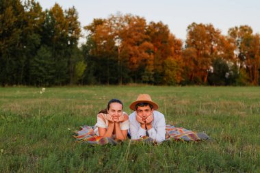 Çimenli bir tarlada battaniyeye uzanmış iki insan, güneş arka planda batarken ağaçlarla çevrili olarak ellerini dinlendiriyorlar..