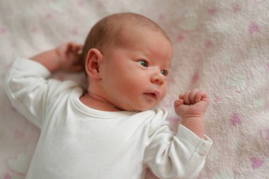 Yeni doğmuş bir bebek, çevreyi izlerken yumuşak bir battaniyenin üzerinde dinlenir. Bebek basit beyaz bir kıyafet giyiyor ve parlak gözlerle merakını gösteriyor..