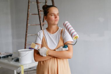 Çapraz kollu bir kadın elinde boya silindiri ve fırçayla odada duruyor. Arka planda bir merdiven ve boya malzemeleri var..