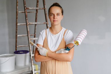 Kolları çapraz duran, elinde boya silindiri tutan ve merdivenli bir odada fırça ve yerde boya kovaları olan bireyler..