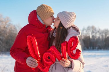 Karda birbirine yakın duran bir çift ellerinde aşk yazan kırmızı bir tabelayla birbirlerini öpüyorlar. Sahne açık bir gökyüzünü ve uzaktaki ağaçları gösteriyor..