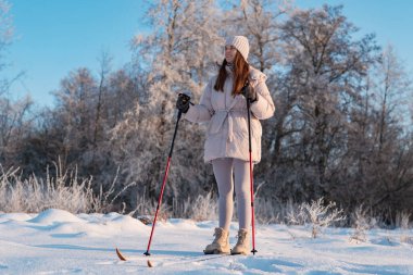 Karlı bir ormanda bir kadın kış etkinliklerinin tadını çıkarırken kayak direklerini kullanıyor. Gökyüzü açık ve ağaçlar donuyor..