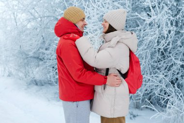 Buzlu ağaçlarla çevrili karlı bir alanda bir çift birbirine yakın duruyor. Gülümserken ve birlikte vakit geçirirken kalın ceket ve şapkalar giyerler..