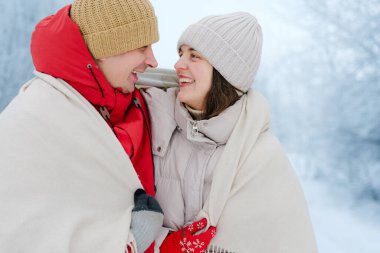 Karlı bir alanda battaniyeye sarılı iki kişi kış gününün tadını çıkarıyor. Birbirlerine gülümsüyorlar ve kalın şapkalar ve ceketler giyiyorlar..