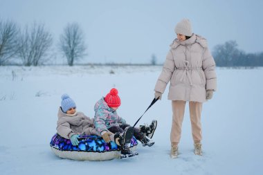 İki çocuk, anneleri ipi tutarken bir kar tüpünde oturuyorlar. Bir kış günü arka planda ağaçlarla karlı bir alanda oynarlar..