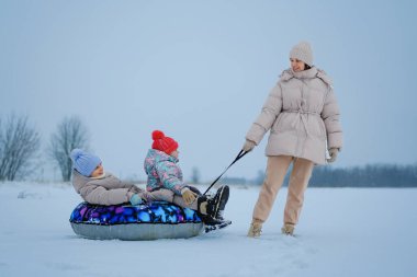 Bir ebeveyn karda şişme bir tüple iki çocuğu çeker. Çocuklar kalın kıyafetler ve şapkalar giyerler. Geniş bir alanda soğuk bir kış günü..