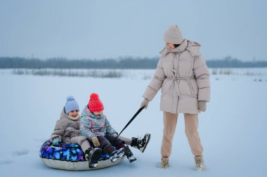 Bir ebeveyn kızağı tutarken iki çocuk bir kızağın üzerinde oturur ve onlara kışın giydikleri karlı arazide rehberlik eder. Soğuk bir gün..