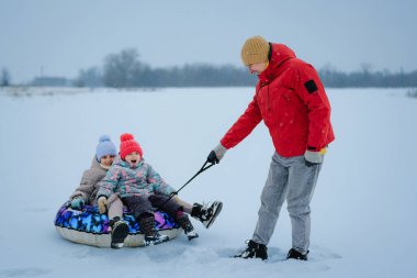 İki çocuk bir kızakta otururken bir kişi onları kış öğleden sonra karlı bir tarlada sürüklüyor. Kar zemini kaplıyor ve arka planda ağaçlar var..