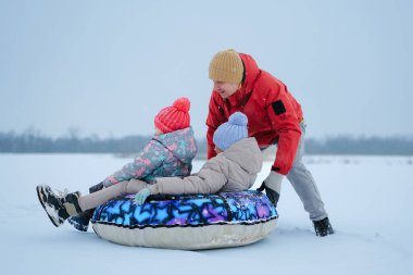 İki çocuk renkli bir metroda otururken bir yetişkin onlara karlı bir alanda yardım ediyor. Sıcak kıyafetler giyerler ve soğuk kış gününün tadını çıkarırlar..