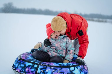 Bir ebeveyn karda renkli bir tüpte otururken çocuğuna sarılır. Kış boyunca birlikte eğleniyorlar ve gülümsüyorlar..