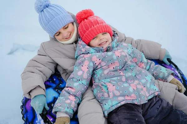İki çocuk karda oynarken bir kızağın üzerinde birlikte otururlar. Kış palto ve şapkalarını takıp kışın dışarıda eğlenirler..