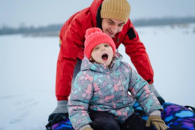 Bir çocuk ve yetişkin karlı bir tepeden kayıyorlar. Çocuk, yetişkin anın tadını çıkarırken heyecanlı görünüyor. Etraflarında kar hafifçe yağıyor.