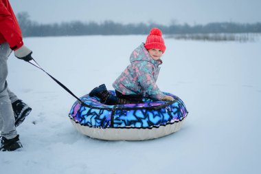 Karlı bir tarlada bir çocuk bir yetişkin tarafından kızağa çekiliyor. Çocuk kış havasında oynarken renkli bir ceket ve parlak bir şapka giyer..