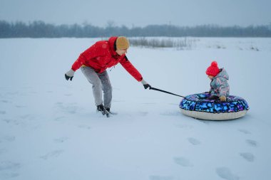 Bir ebeveyn kış boyunca bir çocuğu kar tüpüyle karlı bir tarlada sürüklüyor. Çocuk renkli kışlık giysiler giyiyor ve mutlu bir şekilde gülümsüyor..