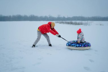 Bir ebeveyn, bir çocuğu karlı bir tarlada kar borusuna çeker. Arazi beyaz karla kaplıdır ve bugün kar yağışı olan bir kış günüdür..