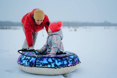 Bir ebeveyn, bir çocuğun karda kar tüneline binmesine yardım eder. Donmuş bir gölde kışlık giysiler giyip karlı havanın tadını çıkarıyorlar..
