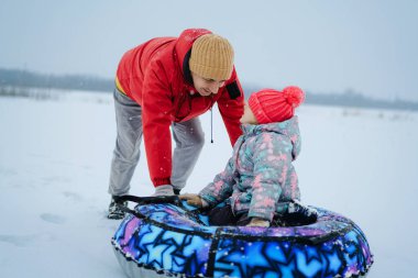 Karlı bir alanda ebeveynleri onlara yardım ederken bir çocuk renkli bir kar borusu çemberinde oturuyor. Etraflarında kar yağıyor. Eğlenceli bir kış sahnesi yaratıyorlar..