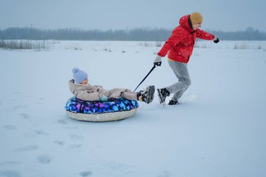 Bir yetişkin, bir çocuğu karlı bir tarlada renkli bir kar tüpüne çeker. Kışın birlikte oynarlarken derin karların içinden geçerler..