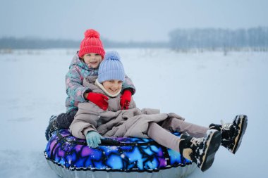 Karlı bir bölgede iki çocuk bir kar tüpünün üzerinde oturuyor. Gülümsüyorlar ve karda eğlenirken kışlık giysiler ve şapkalar giyiyorlar..