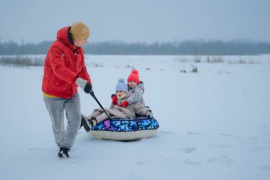 İki çocuk bir kızakta otururken ebeveynleri onları karla kaplı zeminde sürüklüyor. Sahne, karlı bir arazide kış aktivitelerini gösteriyor..