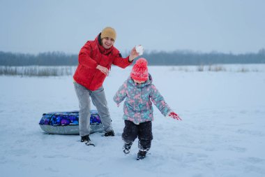 Yetişkinler kartopu atarken bir çocuk karda neşeyle koşuyor. Arka planda bir göl ve ağaçlarla dolu bir kış bölgesindeler..
