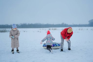 Çocuklar karda vakit geçirirken bir yetişkin, soğuk bir kış günü geniş bir karlı bölgede kızakla onlara yardım ediyor..