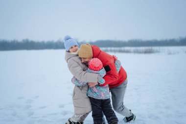 Bir baba karlı bir arazide iki çocuğunu kucaklıyor. Çocuklar karda beraber oynarken gülümsüyor ve kalın kışlık giysiler giyiyorlar..