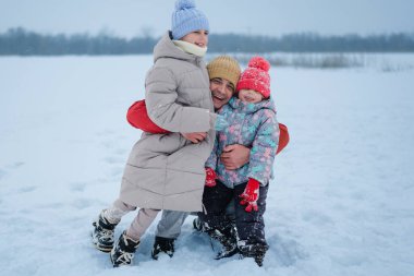 İki çocuk, kış havasının tadını çıkarırken karlı bir tarlada bir yetişkinle birlikte duruyor. Kar yağarken gülümserler ve yüzlerini ve elbiselerini örterler..